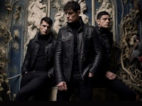 three men in black leather jackets posing in front of an ornate wall